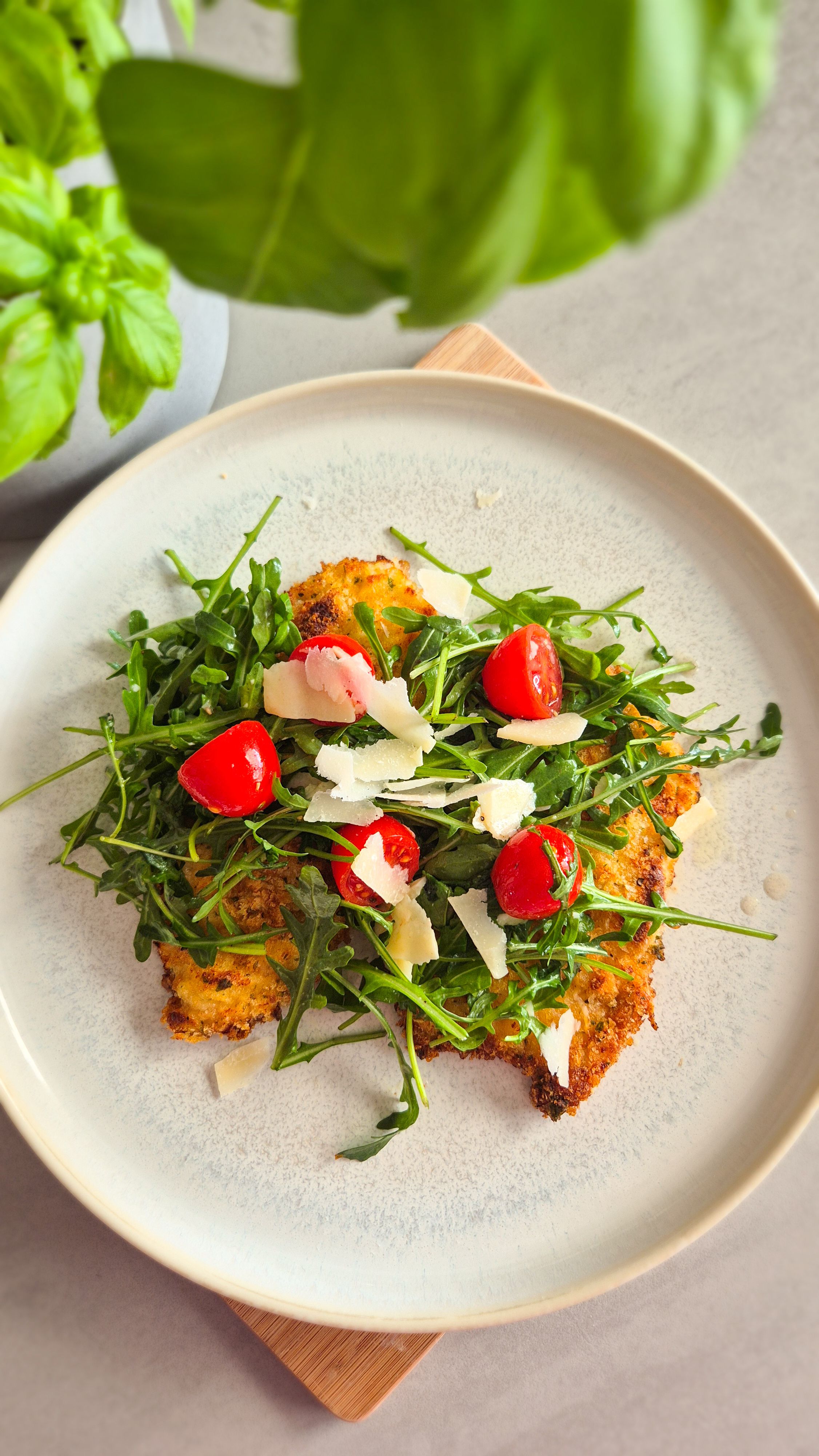 Ein Teller mit knusprig gebackenem Chicken Milanese, belegt mit frischem Rucola, Cherrytomaten und Parmesan-Hobeln, serviert mit einer Zitronenspalte.