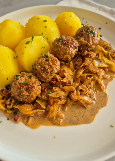 Hackbällchen mit gedünstetem Weißkohl in cremiger Sauce und Salzkartoffeln auf einem Teller.