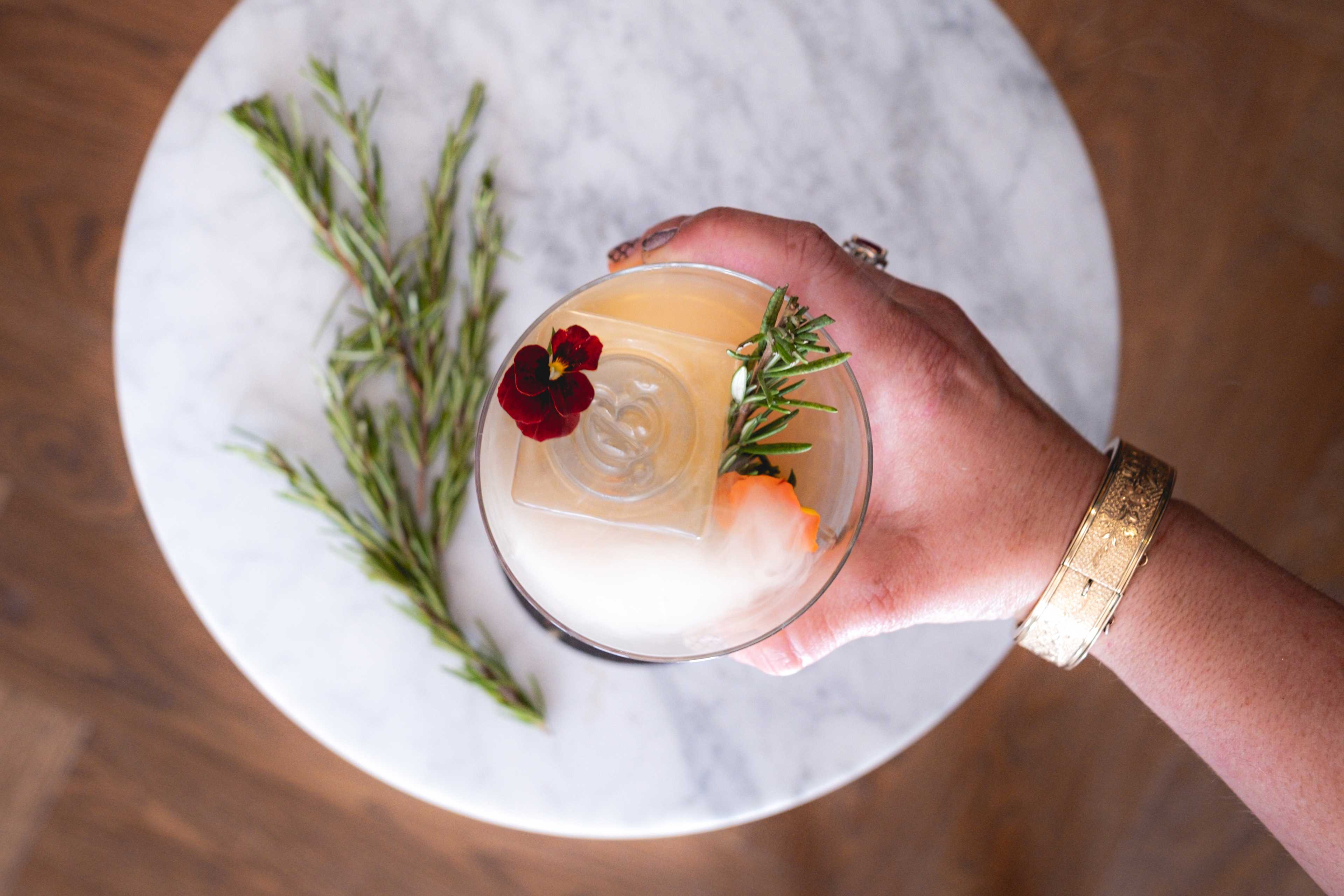 a hand holding a glass of a drink with a sprig of rosemary.