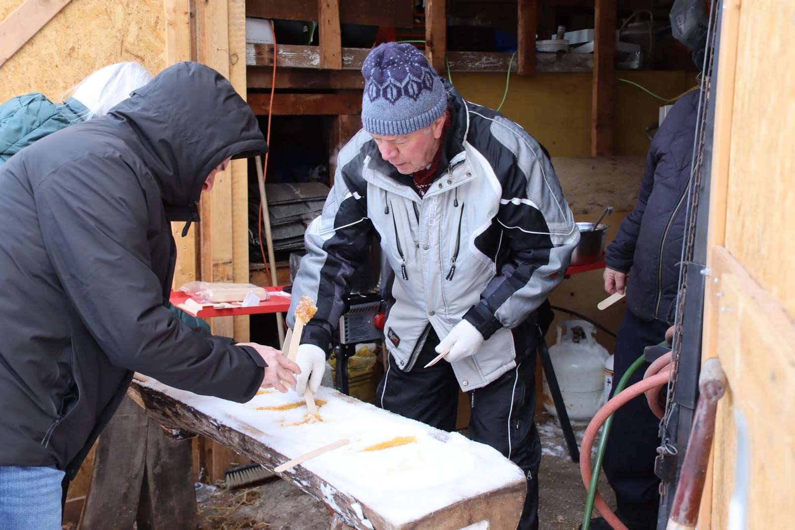 Trois hommes en manteaux d'hiver travaillent autour d'une table en bois recouverte de neige. L'un d'eux utilise un bâton pour ramasser un morceau de tire d'érable sur la neige. Ils portent des gants et des bonnets, et l'environnement est rustique avec des outils et des conteneurs en arrière-plan.