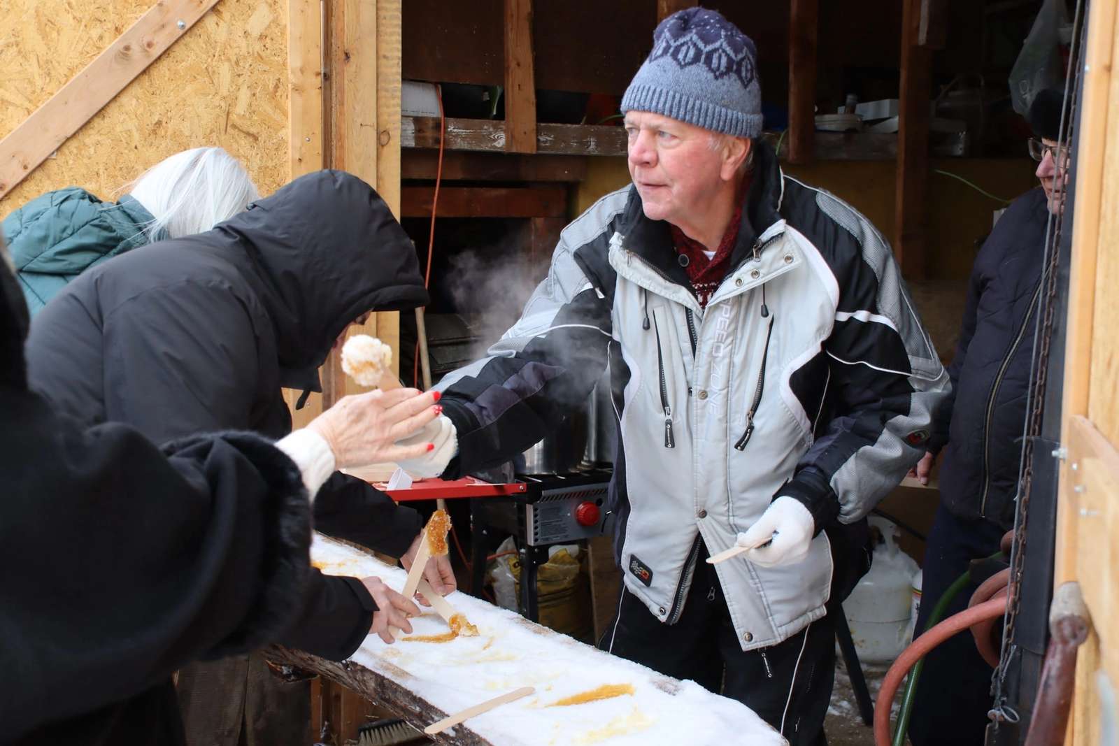 Un homme en manteau et bonnet offre de la tire d'érable sur neige aux invités.
