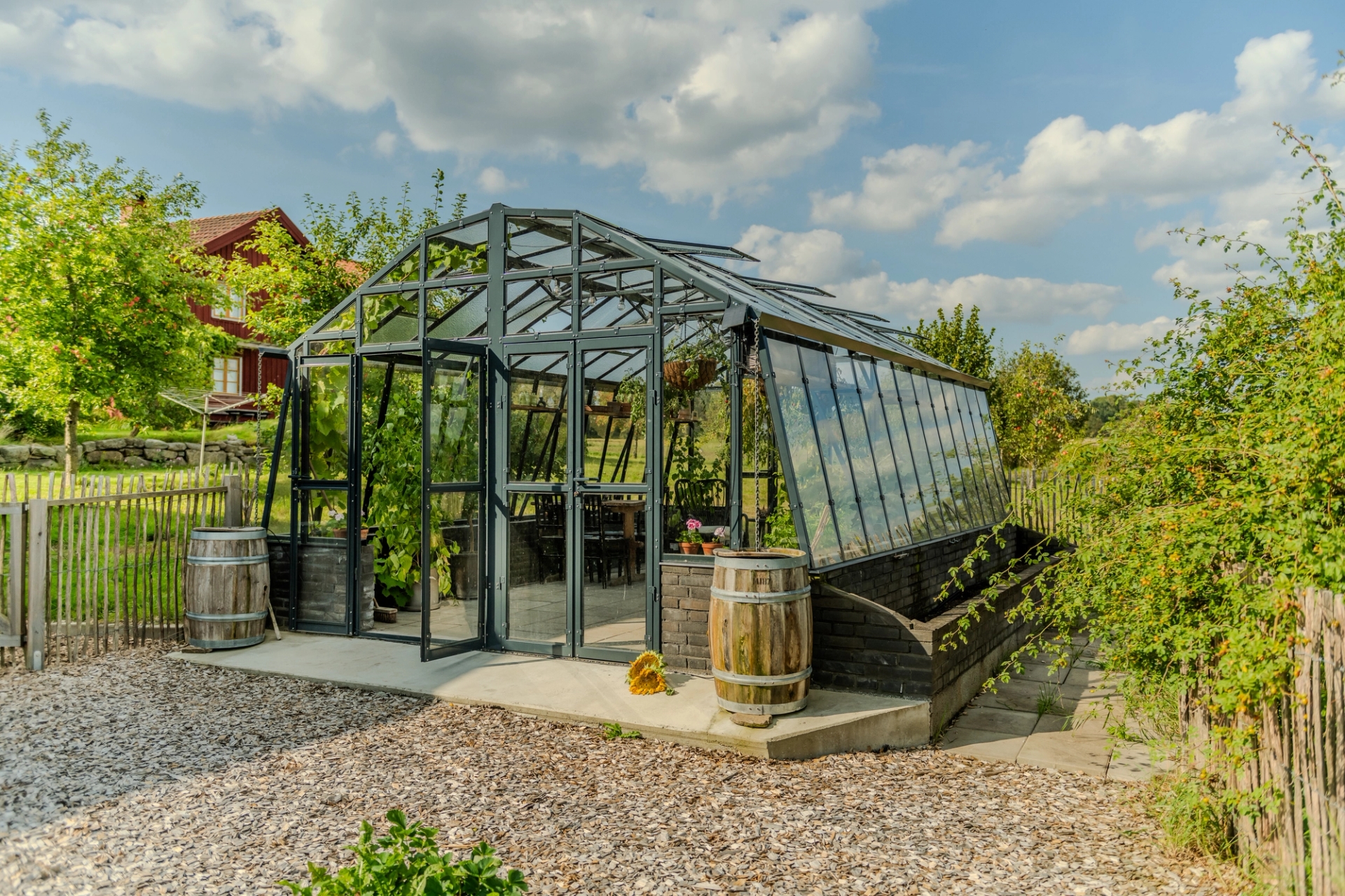 Stormsäkert växthus Variant 426 på mur, med öppen dörr, placerat i en svensk trädgård.