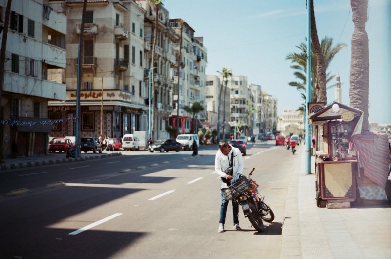 A man wheeling his motorbike across an empty seafront avenue. Echoes of Egypt, photograph by Yannis Papanastasopoulos