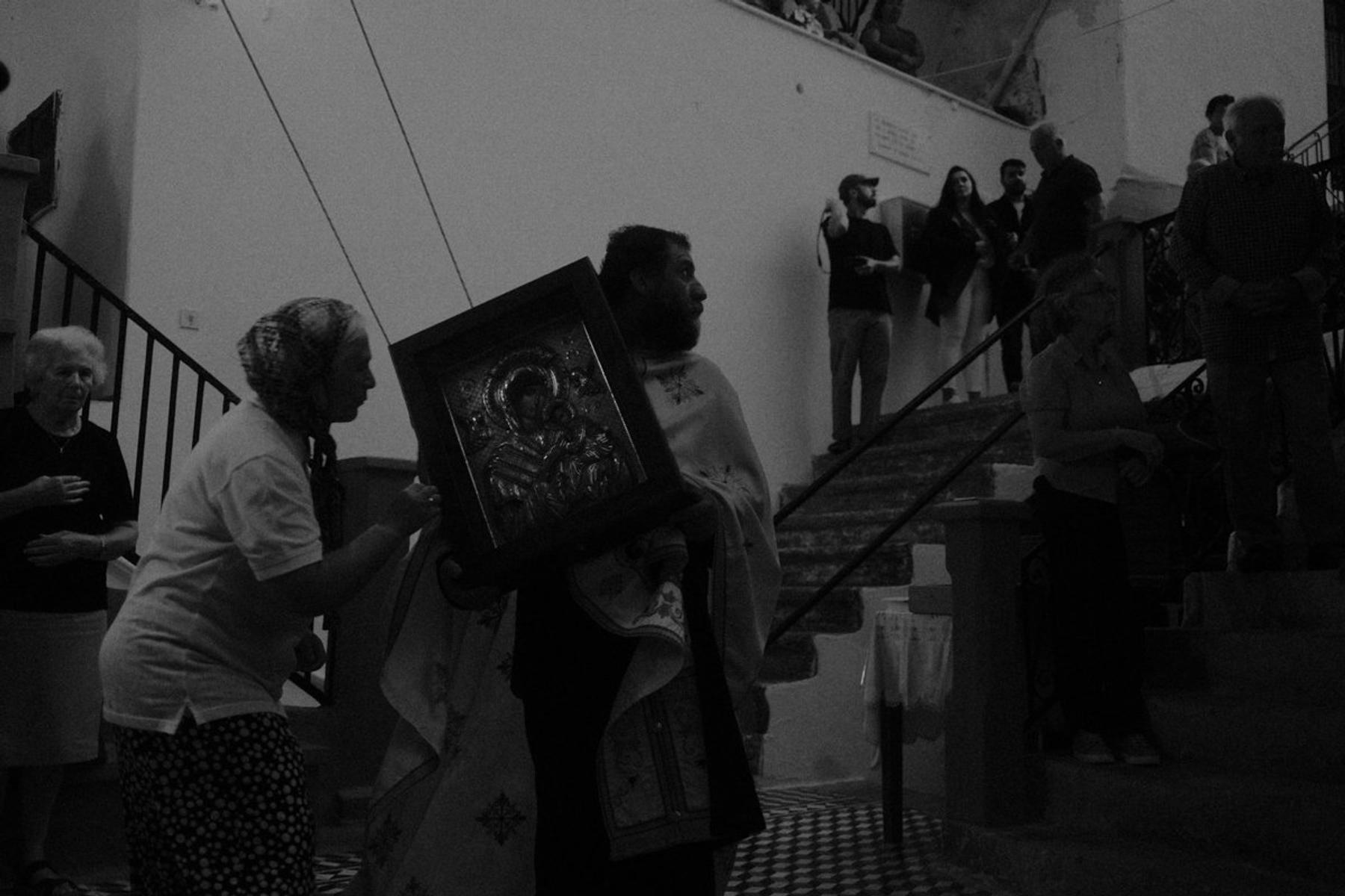 A priest carrying an icon out through a doorway, a woman steadying it beside him