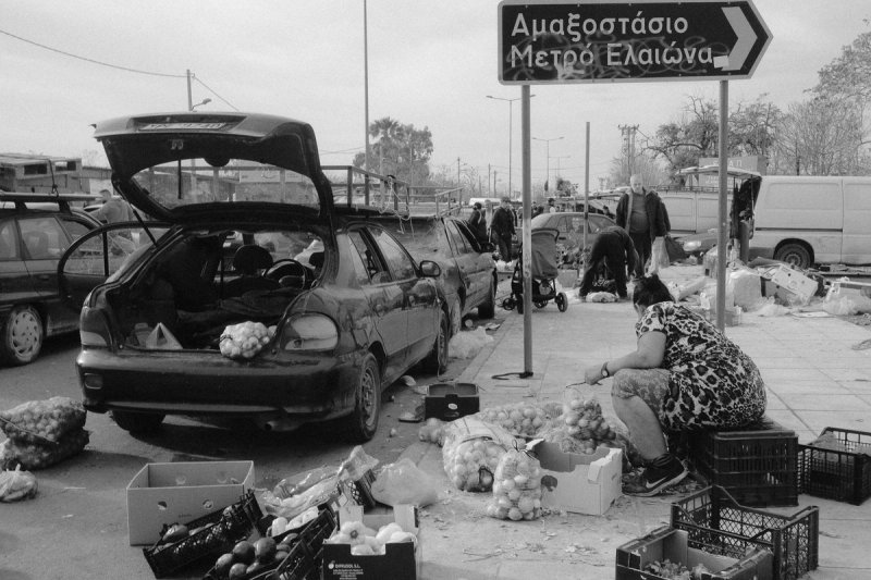A car boot open at the roadside market, a woman bagging onions beneath a metro sign for Elaionas. Elaionas, photograph by Yannis Papanastasopoulos