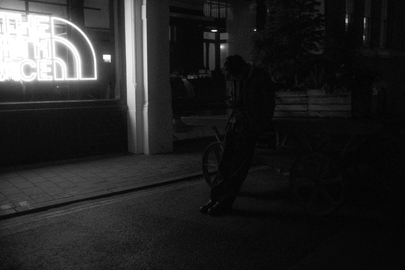 A man bent over a cart on an empty street at night, a neon shopfront sign behind him. London's Untold Stories, photograph by Yannis Papanastasopoulos
