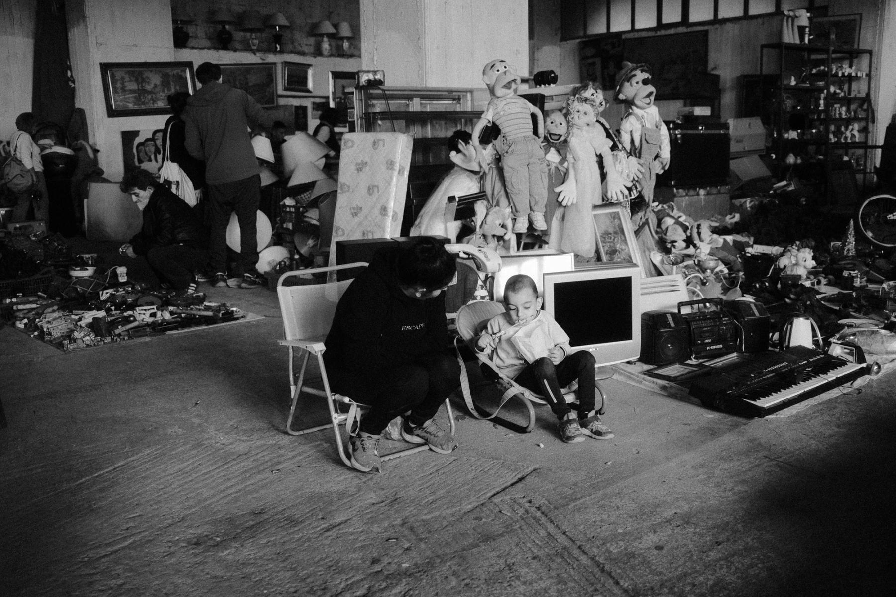 A man crouching beside a toddler on a folding chair, puppets and an old monitor stacked behind them