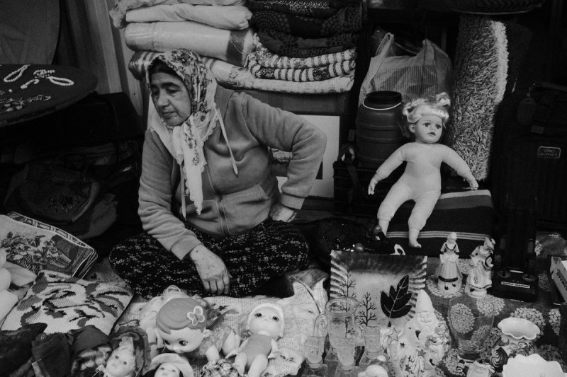 A woman in a headscarf sitting among folded blankets, dolls and figurines laid out in front of her. Elaionas, photograph by Yannis Papanastasopoulos