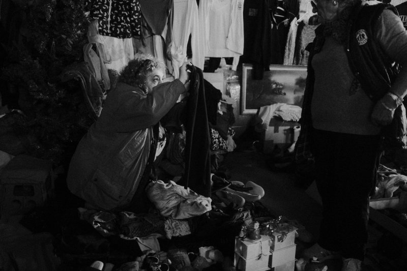A woman kneeling to sort through a heap of clothes while another stands over her. Elaionas, photograph by Yannis Papanastasopoulos