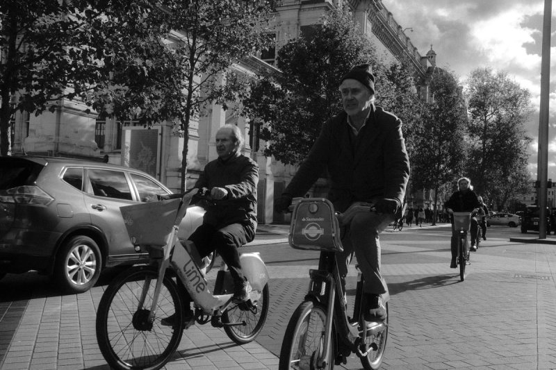 Two older men riding hire bikes along a tree-lined avenue. London's Untold Stories, photograph by Yannis Papanastasopoulos
