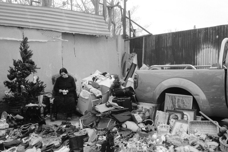A woman sitting alone in an armchair in a yard, shoes and bags spread across the ground. Elaionas, photograph by Yannis Papanastasopoulos