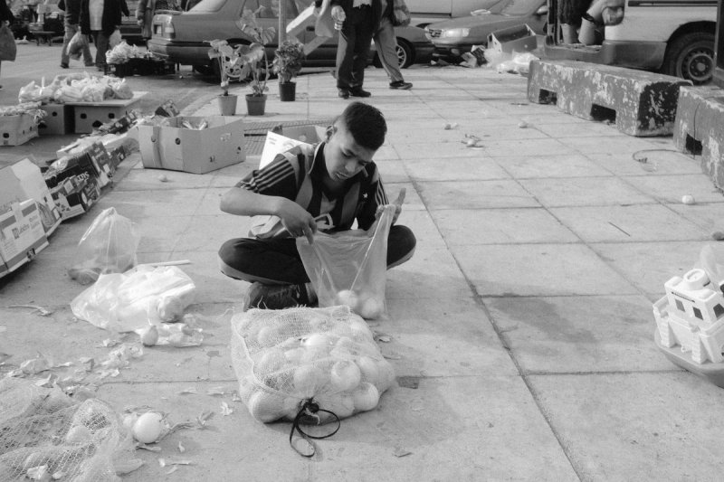 A boy sitting cross-legged on the pavement filling a bag from a sack of onions. Elaionas, photograph by Yannis Papanastasopoulos