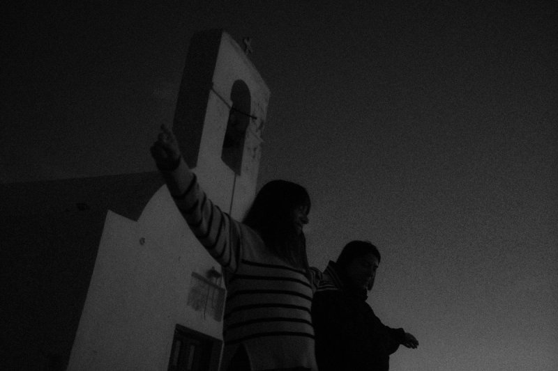A woman reaching up beside a whitewashed bell tower at dusk. Serifos, photograph by Yannis Papanastasopoulos