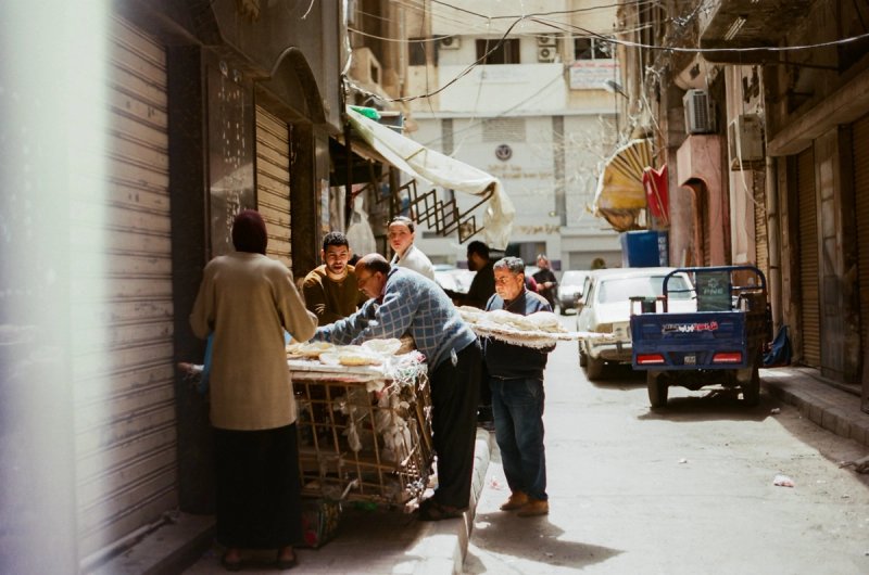 Men crowding around a bread cart in a narrow lane, flatbread stacked along its rail. Echoes of Egypt, photograph by Yannis Papanastasopoulos