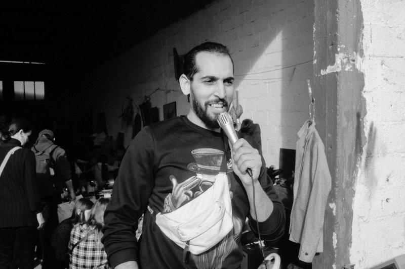 A man laughing into a microphone against a whitewashed brick wall, the market behind him. Elaionas, photograph by Yannis Papanastasopoulos