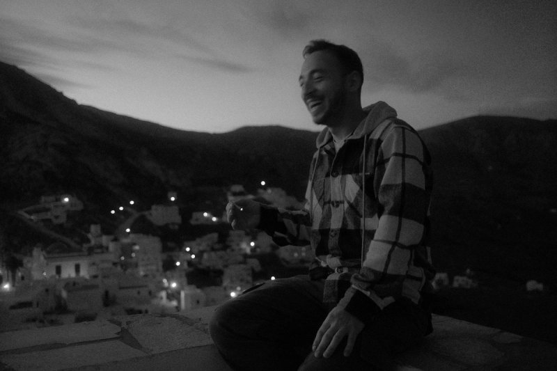 A man laughing on a rooftop at dusk, the village lights coming on below. Serifos, photograph by Yannis Papanastasopoulos