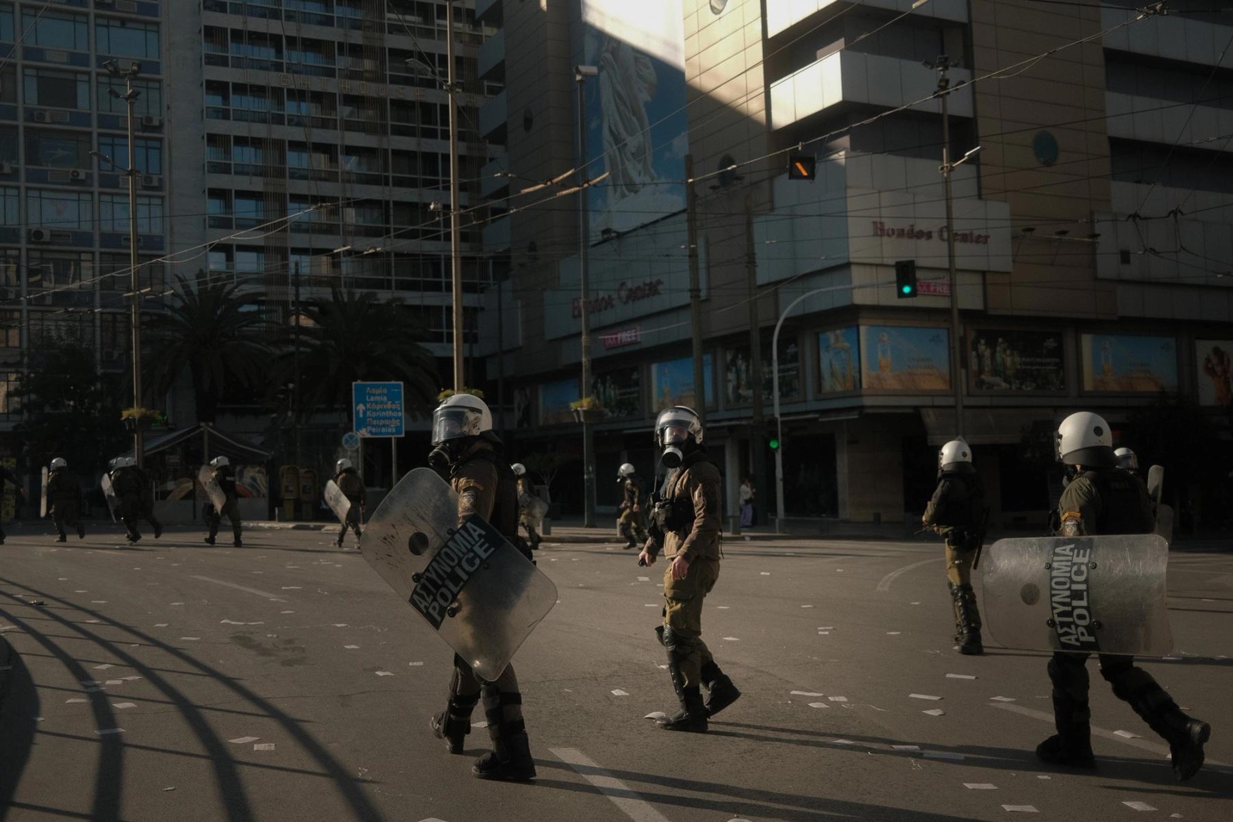 Riot police with shields spread across an empty Athens avenue in low sun