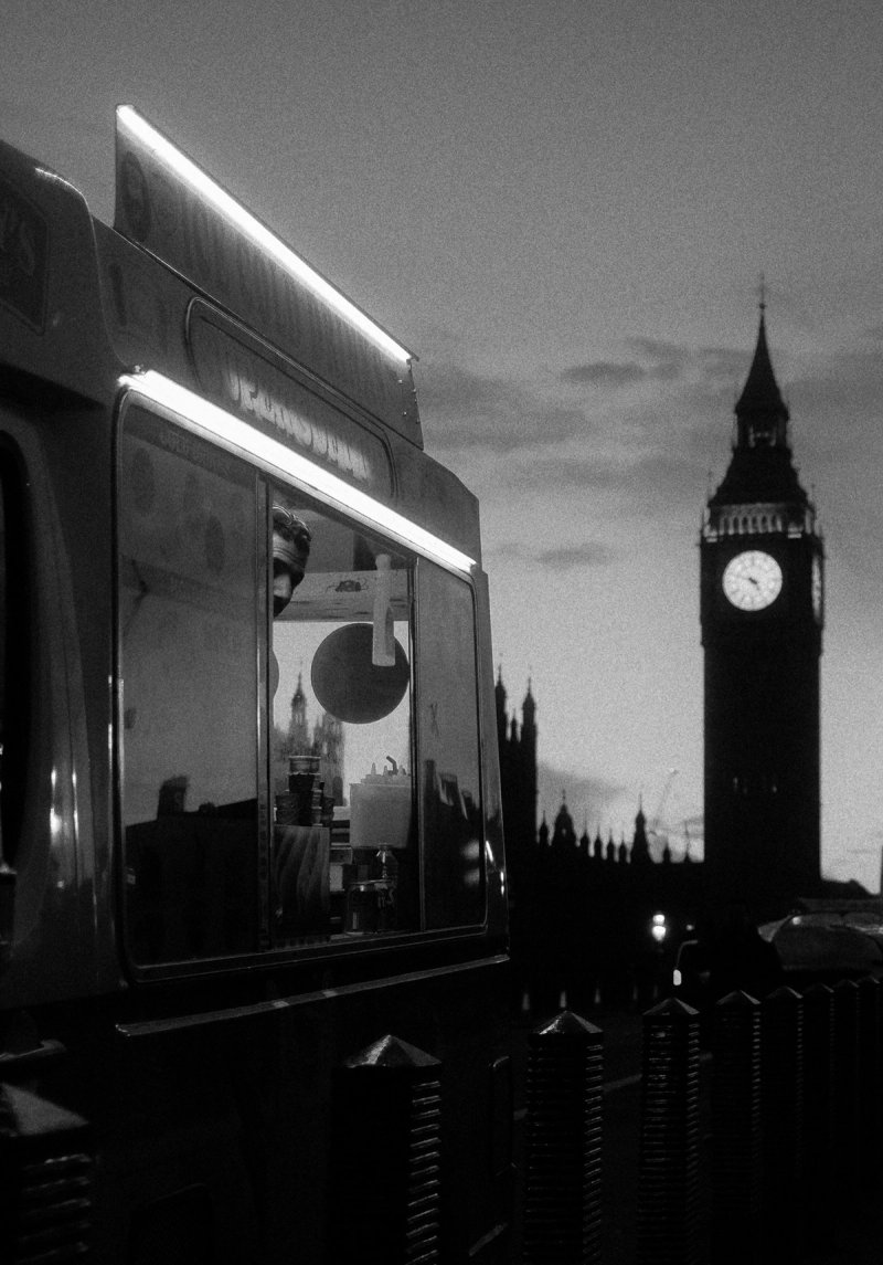 An ice cream van lit up at dusk with Big Ben and Parliament behind it. London's Untold Stories, photograph by Yannis Papanastasopoulos