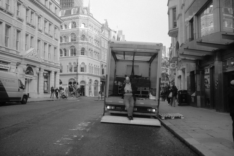 A man standing in the open back of a lorry parked in the middle of a city street. London's Untold Stories, photograph by Yannis Papanastasopoulos