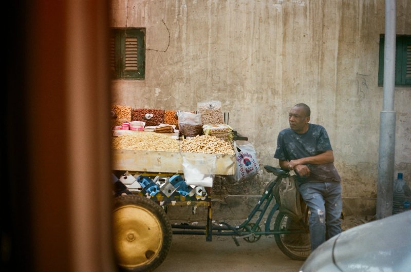A vendor leaning on his nut cart against a bare plaster wall. Echoes of Egypt, photograph by Yannis Papanastasopoulos