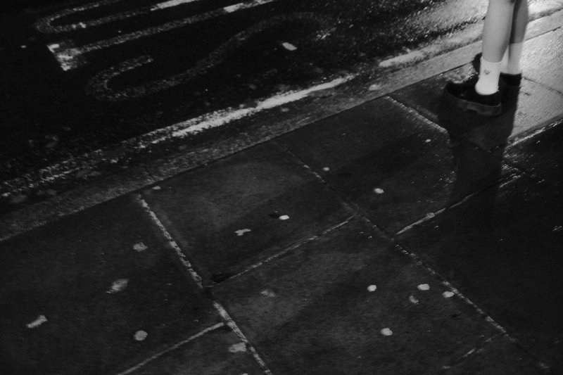 A child's legs and shoes at the edge of a wet pavement at night. London's Untold Stories, photograph by Yannis Papanastasopoulos