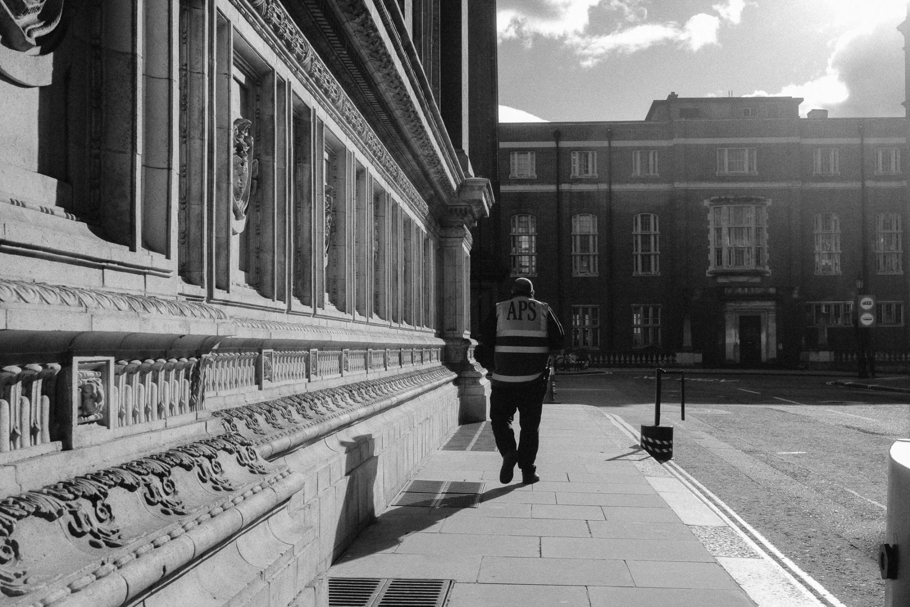 A security guard in a hi-vis vest walking along a carved stone facade in hard morning light