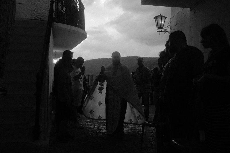 A priest with a censer in a narrow lane at dusk, villagers gathered on both sides. Divine Encounters, photograph by Yannis Papanastasopoulos