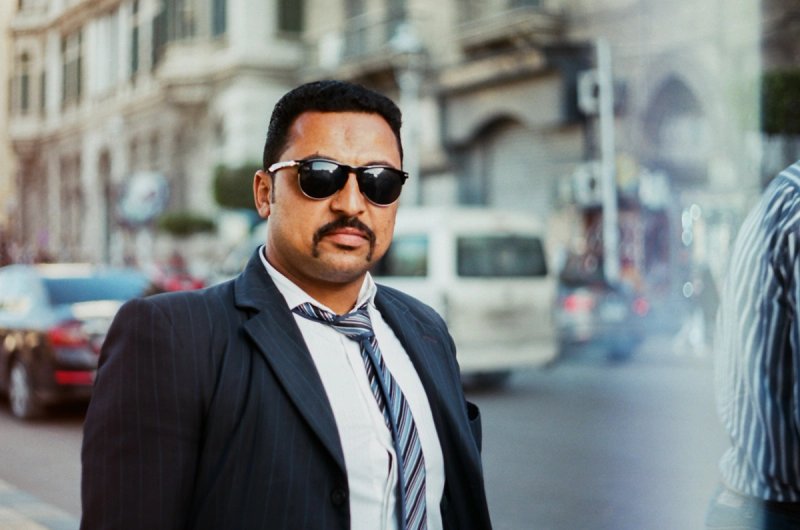A man in a suit and sunglasses on a busy street, turning toward the camera. Echoes of Egypt, photograph by Yannis Papanastasopoulos