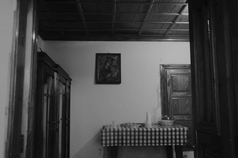 An empty room with an icon on the wall above a table in checked oilcloth. Divine Encounters, photograph by Yannis Papanastasopoulos