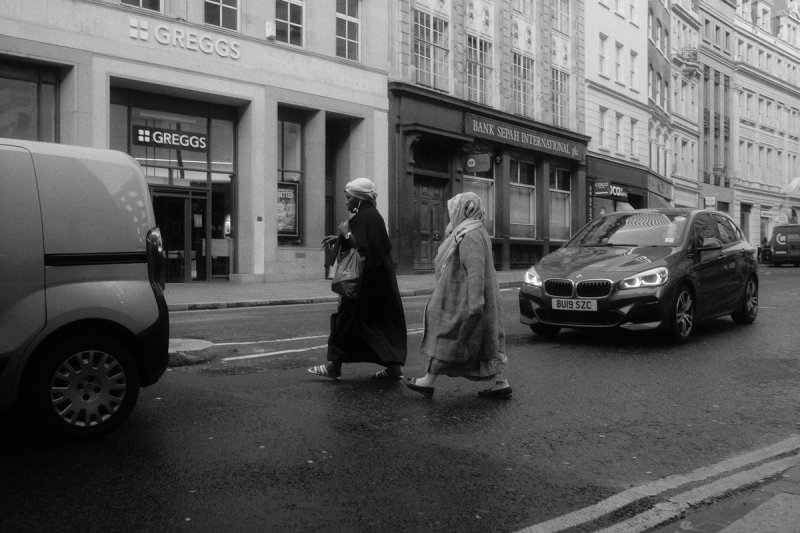 Two women in long coats crossing the road between a van and a waiting car. London's Untold Stories, photograph by Yannis Papanastasopoulos