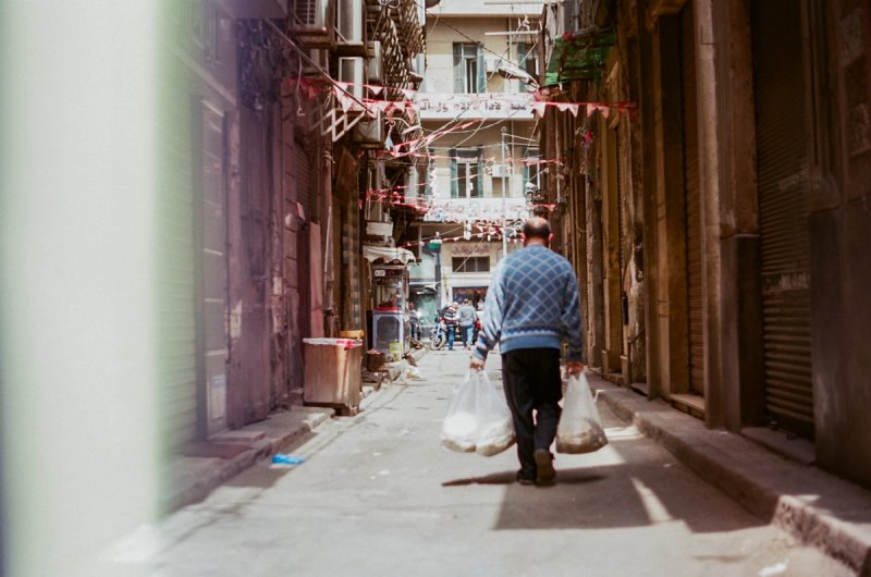 A man carrying two full shopping bags down a lane hung with bunting. Echoes of Egypt, photograph by Yannis Papanastasopoulos