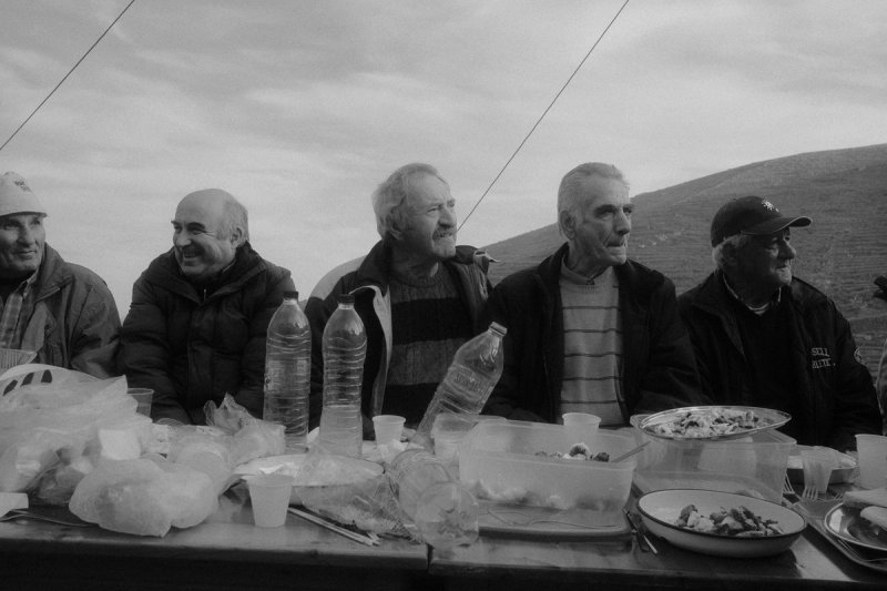 Five men along one table outdoors, plates and water bottles between them. Serifos, photograph by Yannis Papanastasopoulos