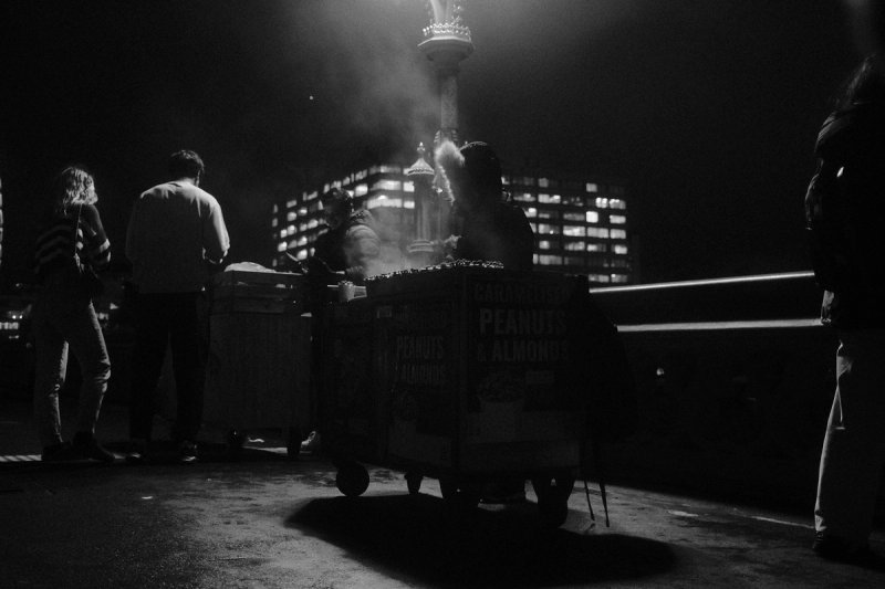 A roasted-nut cart steaming under a lamp at night, office windows lit behind. London's Untold Stories, photograph by Yannis Papanastasopoulos