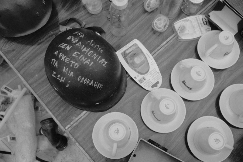 A helmet with Greek writing chalked on it, laid on a table among espresso cups. Elaionas, photograph by Yannis Papanastasopoulos