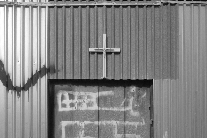 A crucifix mounted on a corrugated metal wall above a spray-painted door. Elaionas, photograph by Yannis Papanastasopoulos