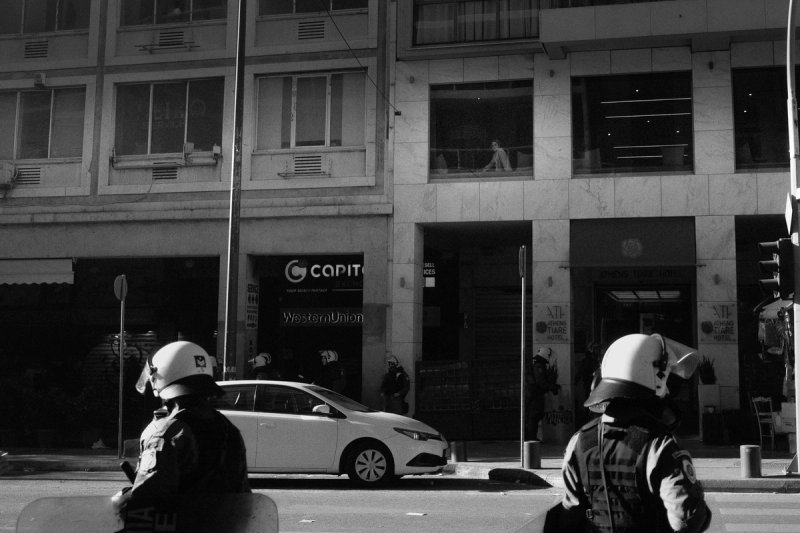 Two helmeted officers waiting at a kerb, a shuttered shopfront across the street. Clashing Shadows, photograph by Yannis Papanastasopoulos