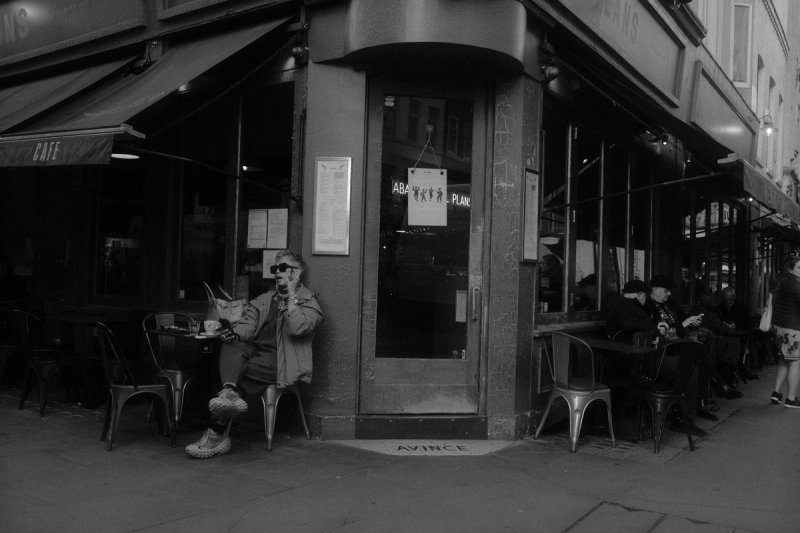 A man sitting alone outside a corner cafe with a coffee, other tables full beside him. London's Untold Stories, photograph by Yannis Papanastasopoulos