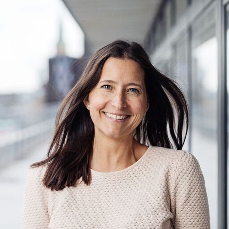 Smiling woman with long brown hair in a light sweater, outdoors.