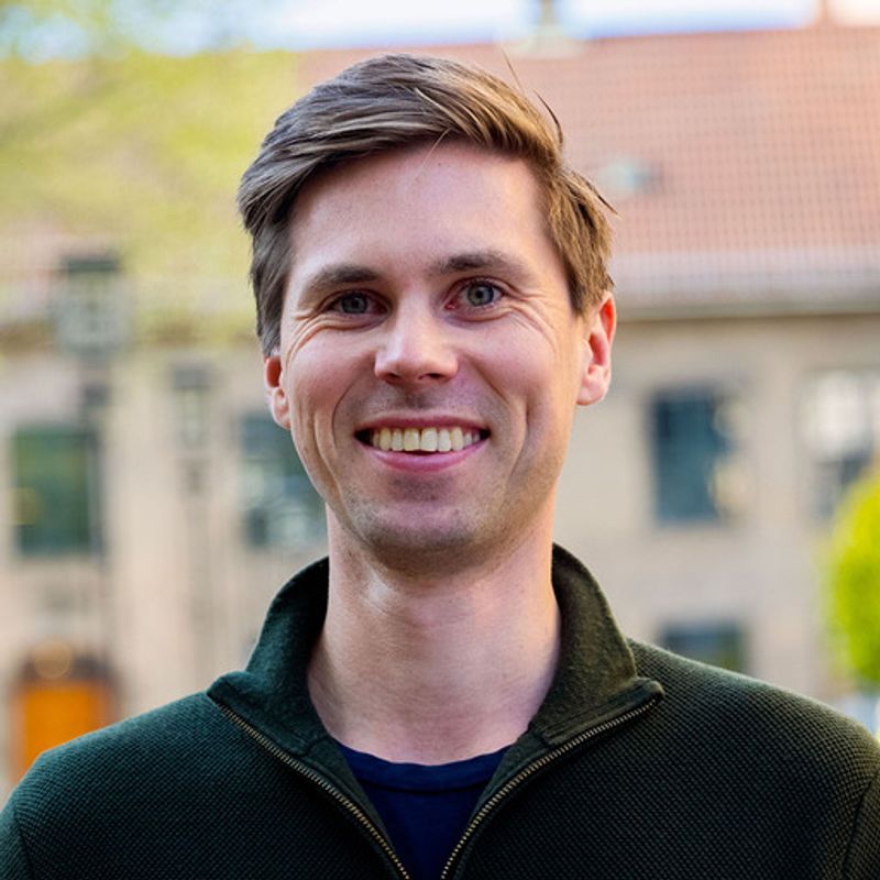 Headshot of a smiling man with light brown hair and blue eyes, wearing a dark green jacket.
