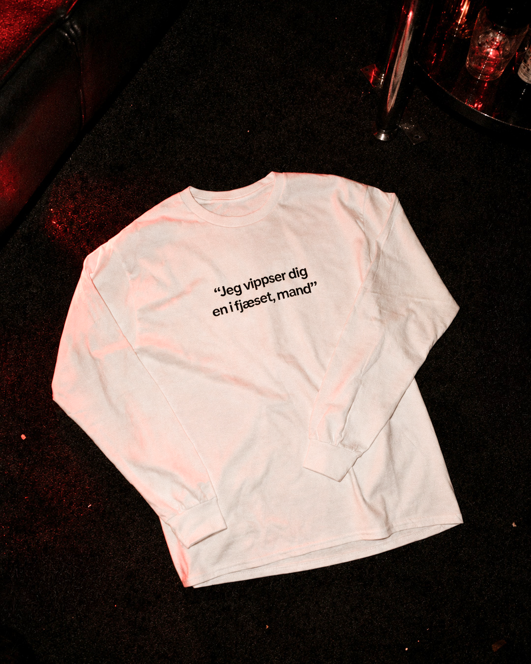 Hvid langærmet T-shirt med teksten “Jeg vippser dig en i fjæset, mand” på en mørk baggrund med rødt lys.