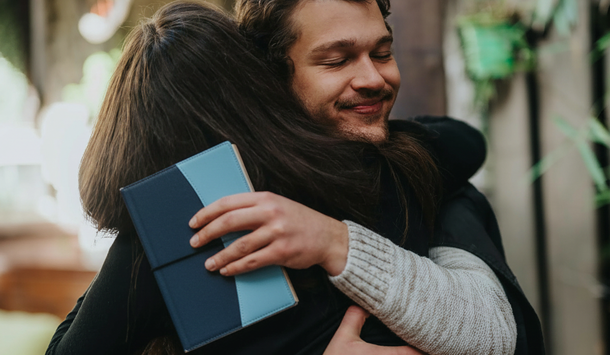 A smiling man with closed eyes hugs another person who holds a two-toned blue book.