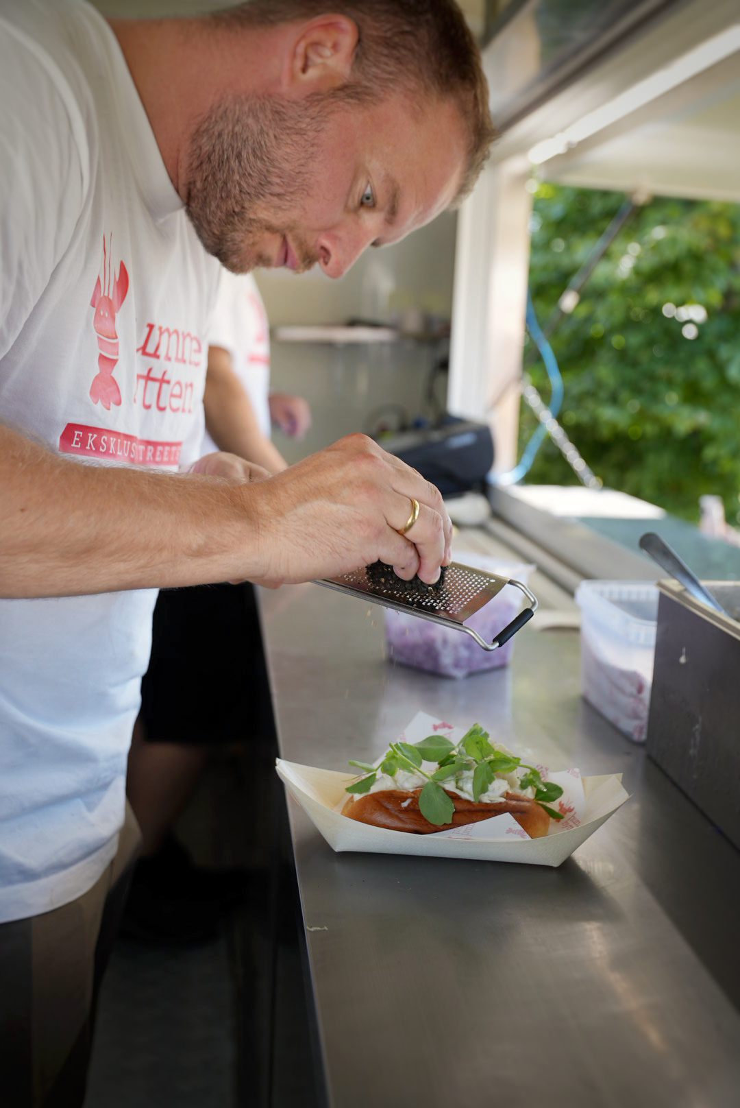 A man grates an ingredient onto a sandwich in a paper boat.