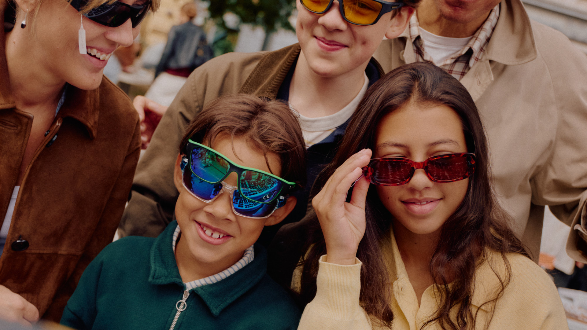 A smiling boy wearing stacked reflective sunglasses and a girl adjusting her red sunglasses, with adults in the background also wearing sunglasses.