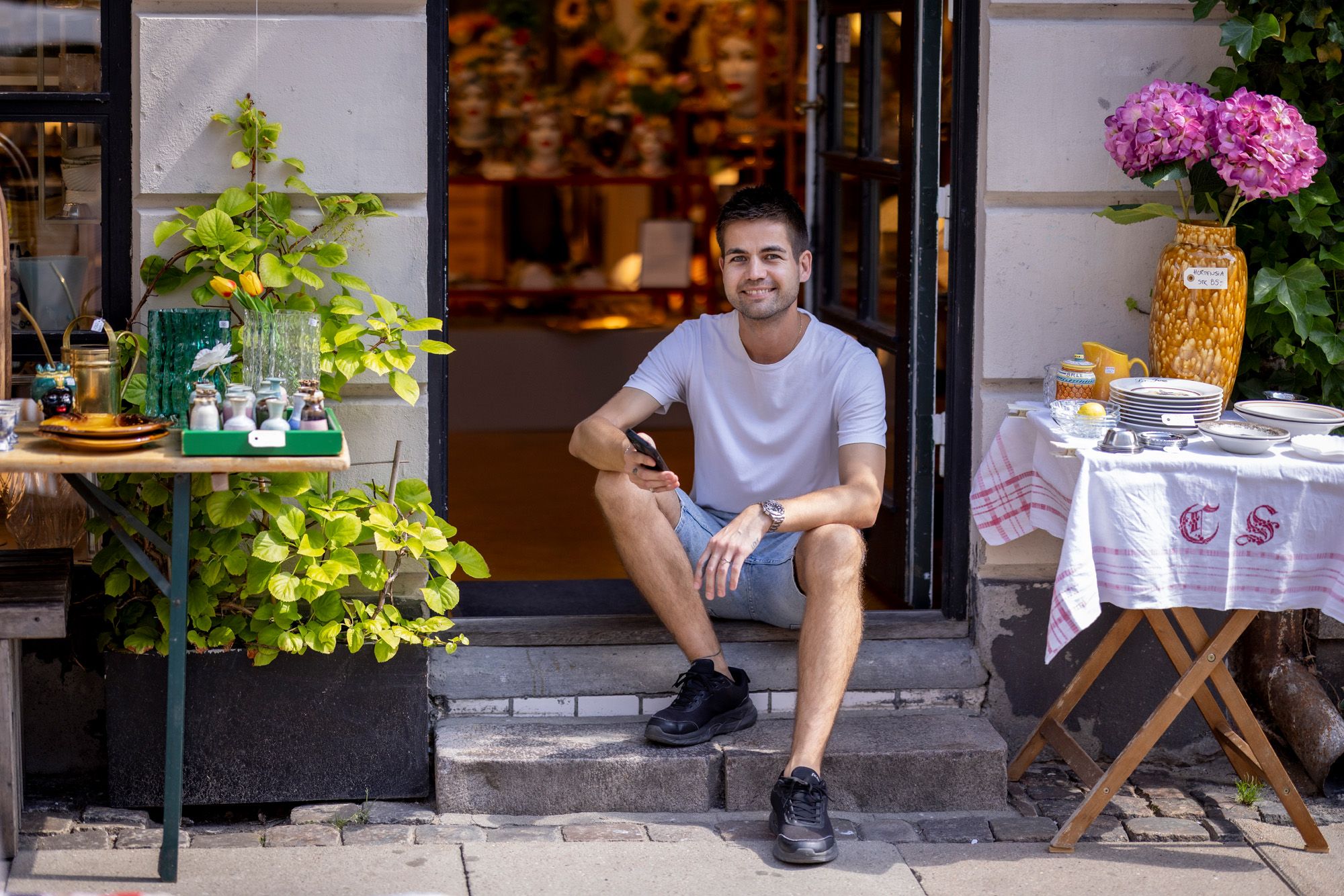 A smiling man sits in a shop doorway, surrounded by outdoor displays of antiques, plants, and flowers.