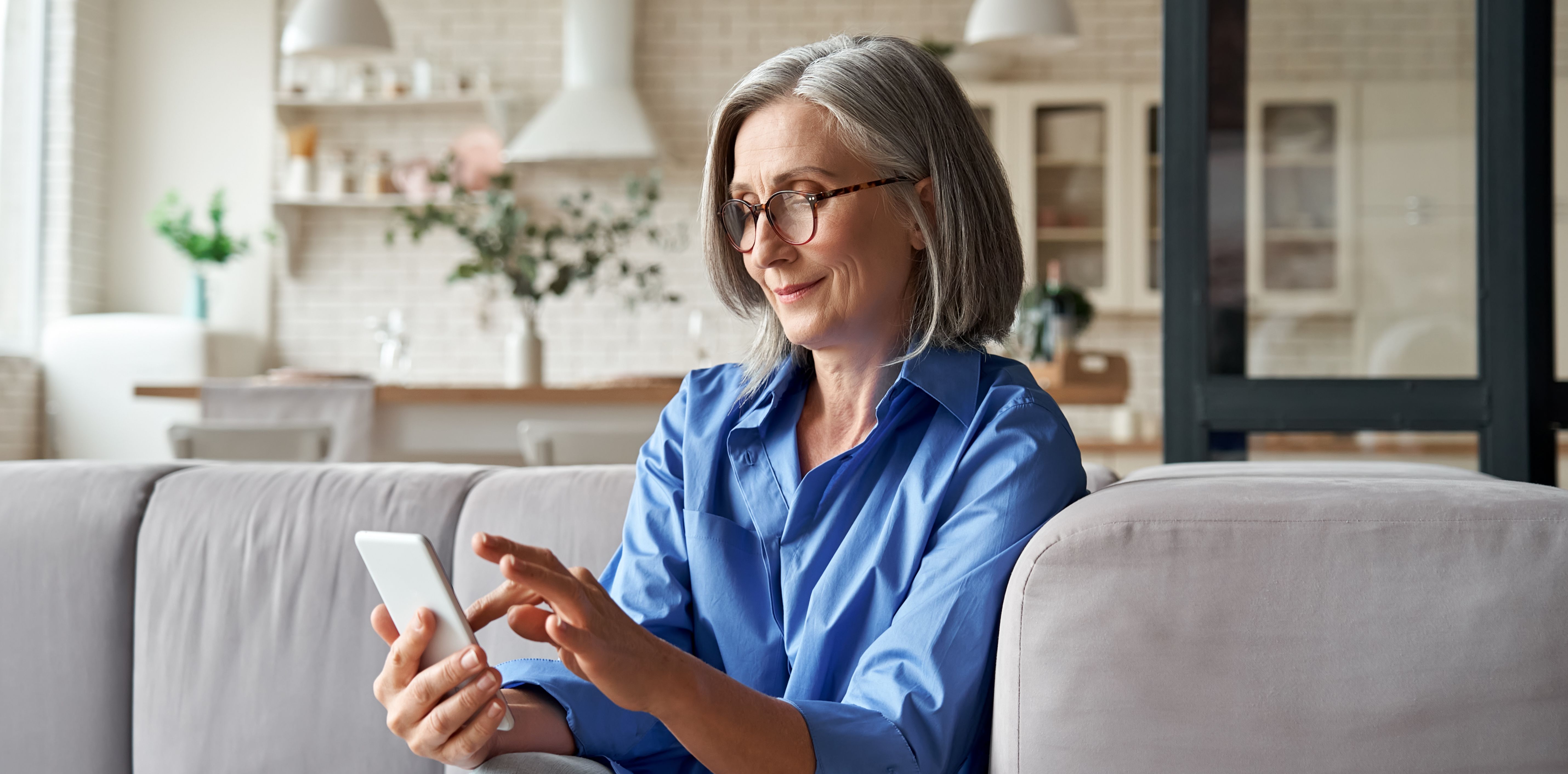 A smiling older woman with grey hair and glasses sits on a sofa, using a smartphone.