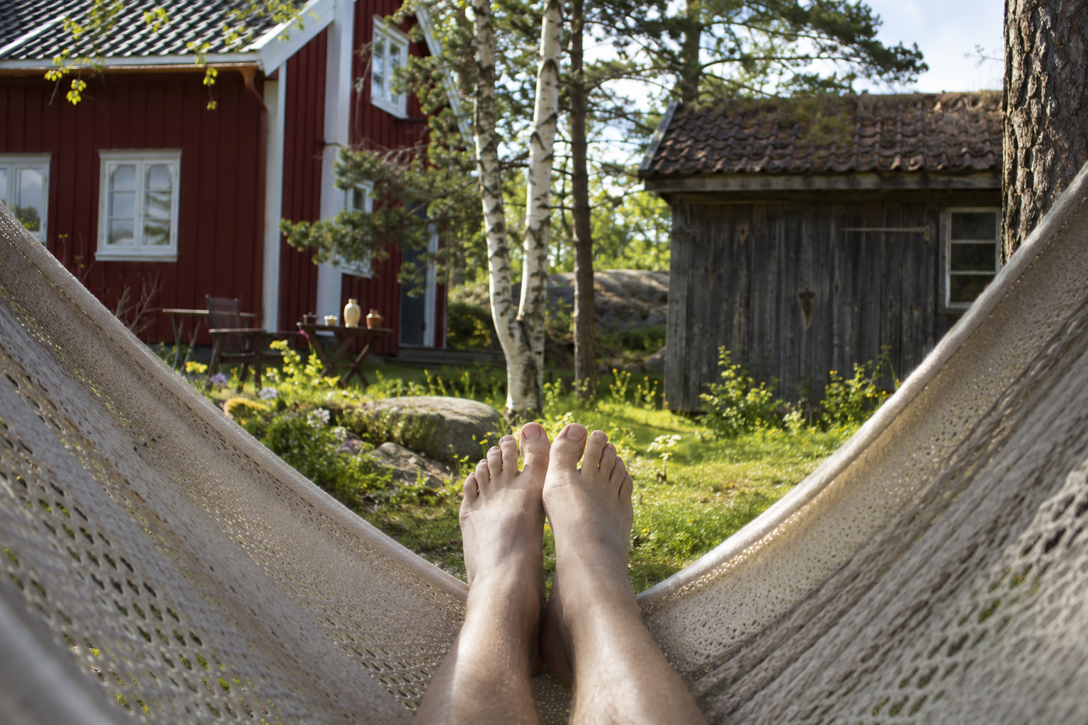 Person's bare feet relaxing in a hammock, overlooking a red house and a rustic wooden cabin.
