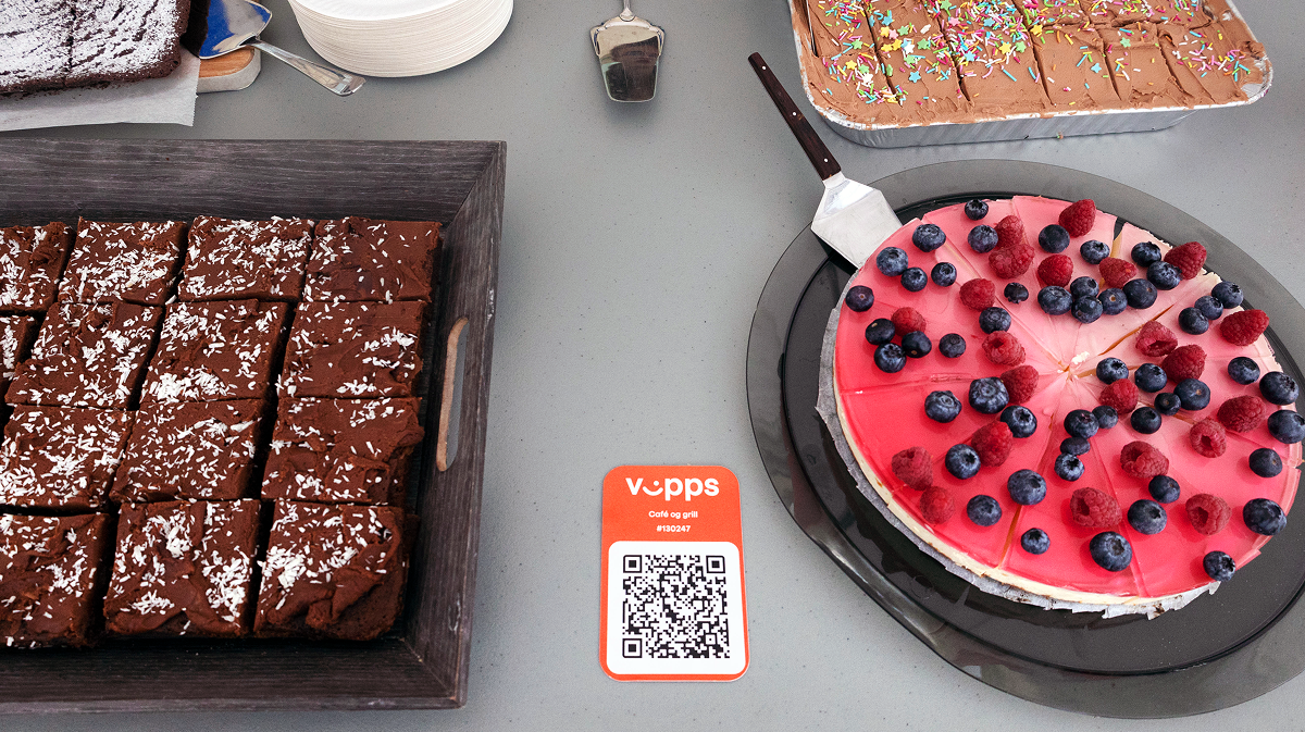 A table with a tray of brownies, a berry-topped cake, another cake with sprinkles, and a Vipps QR code.