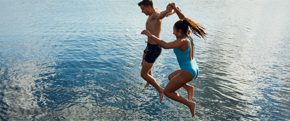 Two young people, holding hands, jump into blue water.