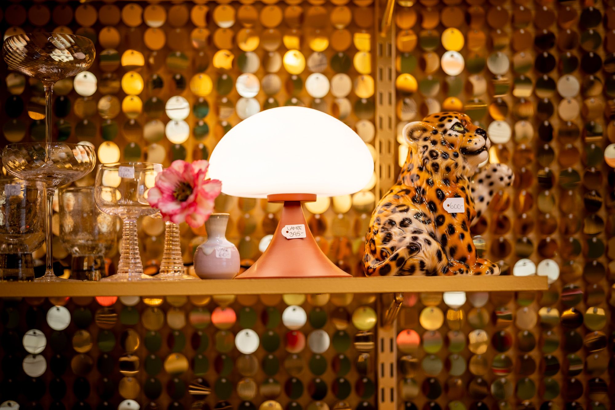 A glowing mushroom lamp, glass stemware, a pink flower, and a leopard figurine on a shelf against a shimmering gold sequin wall.