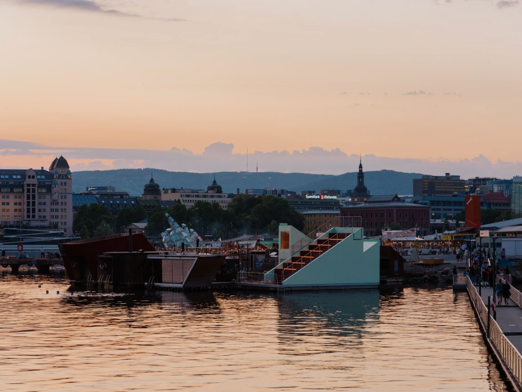 Sunset over a city harbor with floating structures and a distant skyline.